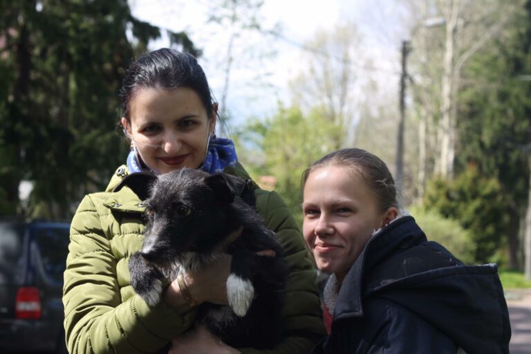 Две женщины улыбаются на улице, одна держит чёрно-белую собаку.