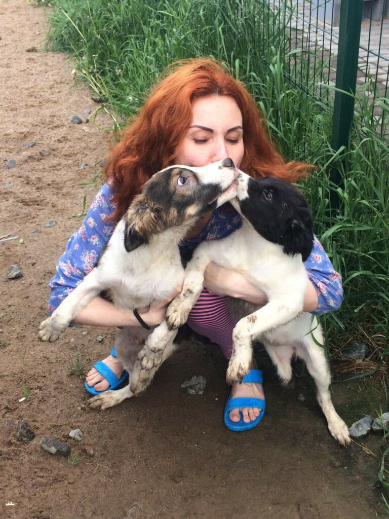 Девушка обнимает двух щенков из приюта — один лижет ей лицо. Снимок на улице: трава, земляная дорожка.