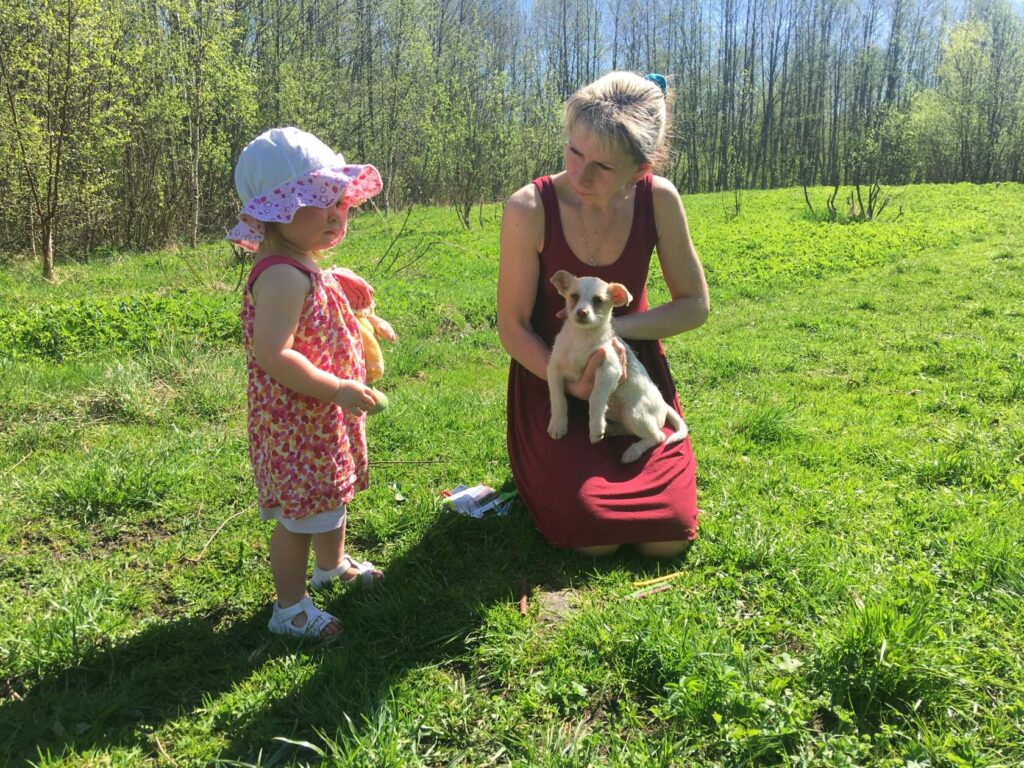 Женщина с щенком на руках и маленькая девочка в панамке на зелёной поляне в солнечный день; день открытых дверей в приюте